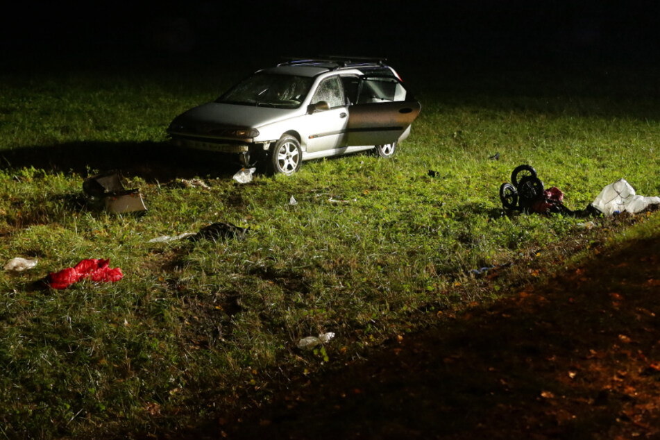Das Unfall-Auto steht neben der Straße auf einer Wiese. Daneben liegen Wrackteile und auch ein Kinderwagen.