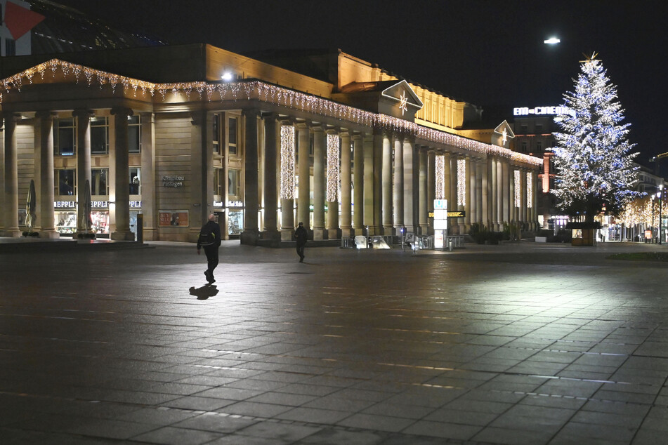 Zwei Männer gehen während der nächtlichen Ausgangssperre über den menschenleeren Schlossplatz in Stuttgart, im Hintergrund ist der Königsbau zu sehen.