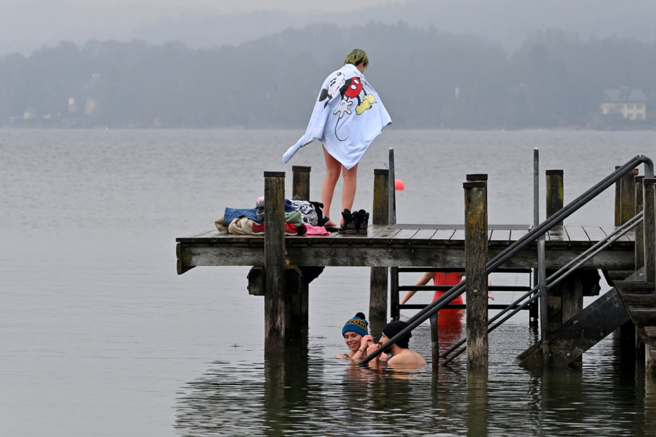 Ein Pärchen mit Pudelmützen sitzt am Steg im Wasser, während zwei Frauen sich auf ihr Bad im Starnberger See vorbereiten.