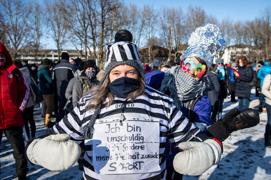 Eine Frau trägt auf der Demo in Rosenheim einen Sträflingsanzug mit einem Schild mit der Aufschrift "Ich bin unschuldig - Ich fordere meine Freiheit zurück. Sofort", während im Hintergrund eine Frau mit einem Aluhut zu sehen ist.
