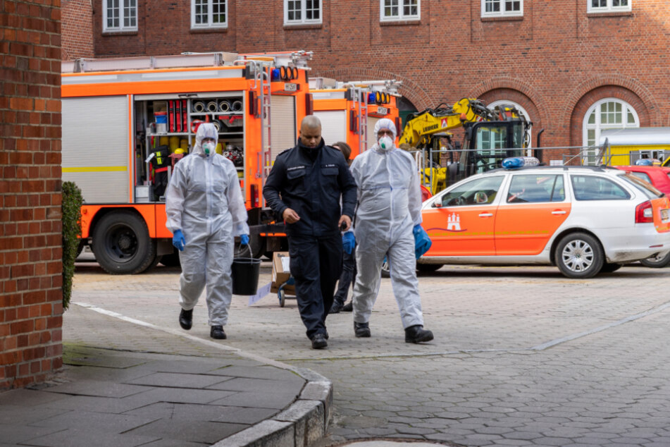 Zwei Mitarbeiter in Schutzanzügen folgen dem Sicherheitspersonal in der Feuerwache.