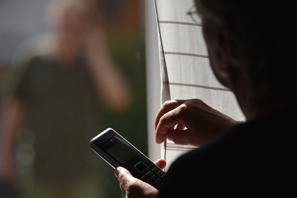 Eine Frau steht mit einem Telefon am Fenster, vor dem ein Mann sie beobachtet (gestellte Szene).