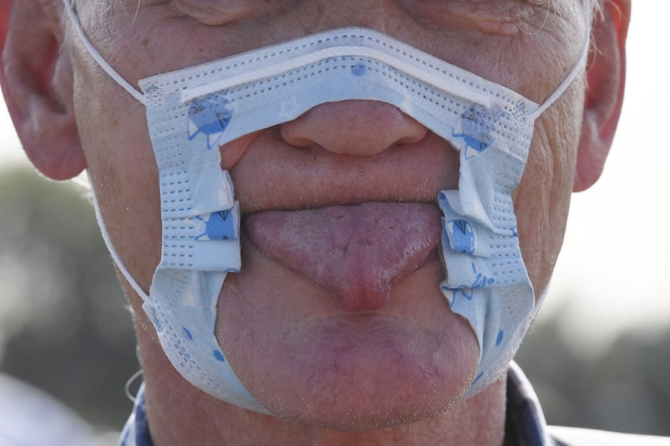 Ein Teilnehmer einer Demonstration protestiert auf den Rheinwiesen mit einer zerschnittenen Maske und herausgestreckter Zunge gegen die Maßnahmen zur Bekämpfung des Coronavirus.