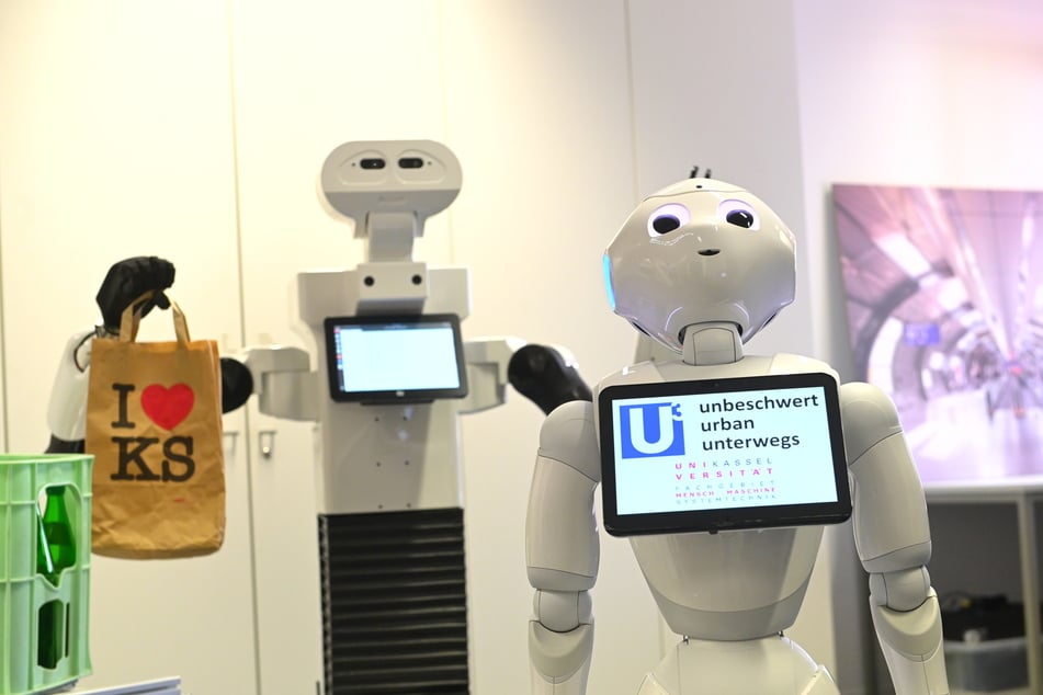 Roboter Pepper (r.) steht vor einem Transportroboter mit Tüte in der Hand.