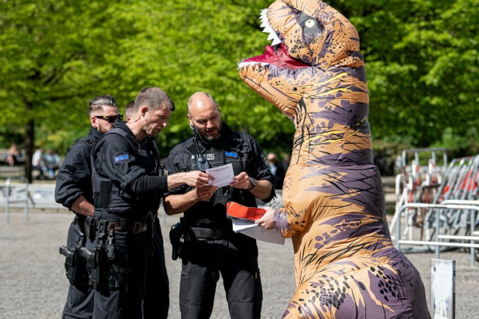 Polizisten sprechen auf der Demonstration gegen die Hygienedemo auf dem Rosa-Luxemburg-Platz mit einem Aktivisten in einem Dinosaurierkostüm.
