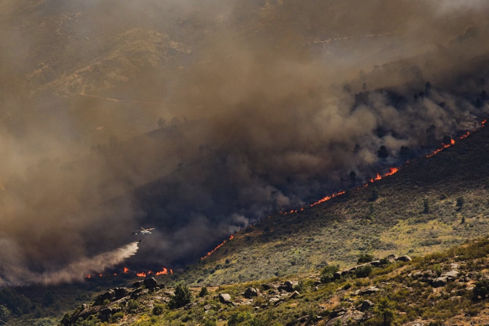 Ein Löschhubschrauber fliegt über einer dichten Rauchsäule während eines Brandes an einem Hang.