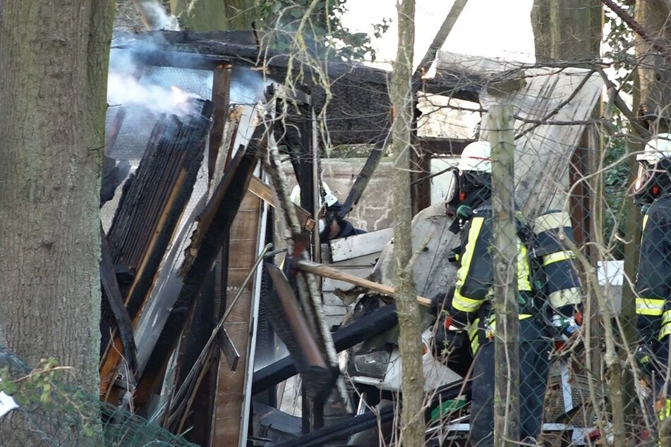 Feuerwehrleute stehen vor der abgebrannten Gartenhütte in Krefeld-Fischeln. Am Samstagabend hatte die Polizei dort bei einem Brand eine Leiche entdeckt.