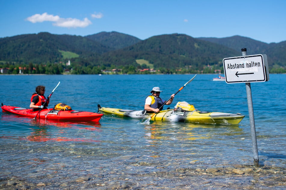 Kajakfahrer paddeln auf dem Tegernsee an einem Schild mit der Aufschrift "Abstand halten" vorbei.