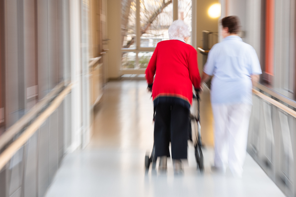 Eine Pflegefachkraft begleitet eine Seniorin am Gehfrei über den Flur eines Seniorenheims.