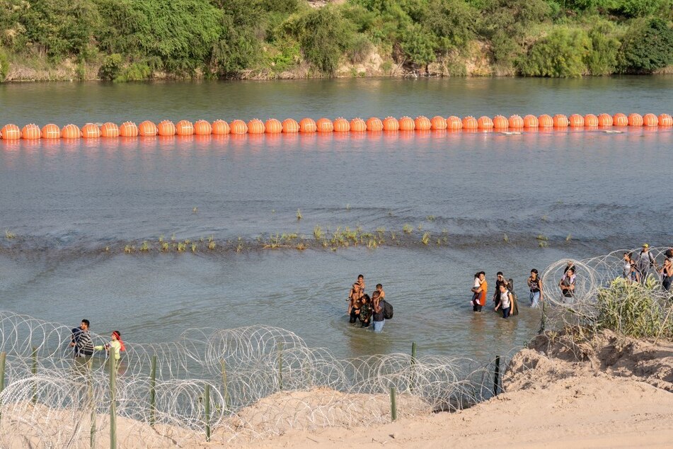 Migrants walk by a string of buoys placed on the water along the Rio Grande border with Mexico in Eagle Pass, Texas, on July 16, 2023.