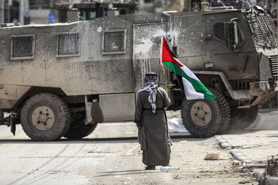 Ein älterer palästinensischer Mann steht mit einer palästinensischen Flagge in der Hand vor einem israelischen Militärfahrzeug. (Archivbild)