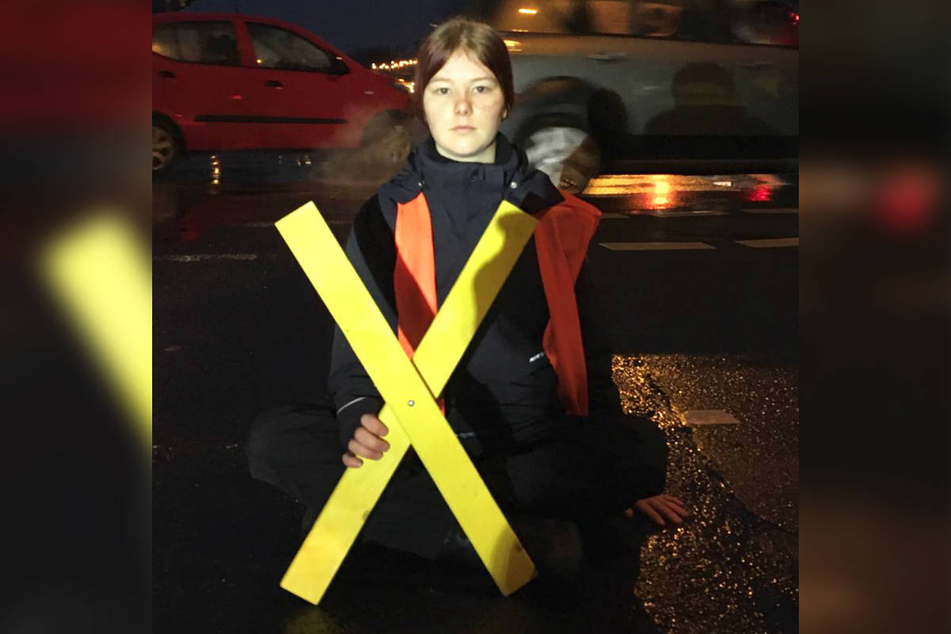 Eine Aktivistin der "Letzten Generation" hat sich am Heckerdamm mit einer Hand auf den Asphalt geklebt, in der anderen hält sie ein gelbes Holzkreuz.