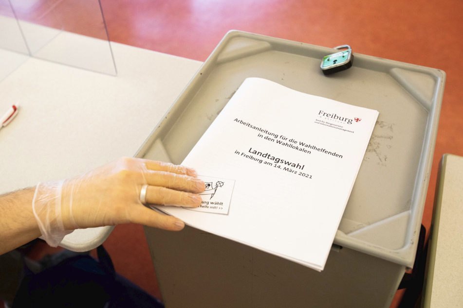 in Wahlhelfer hält die Abdeckung einer Wahlurne in der behandschuhten Hand.