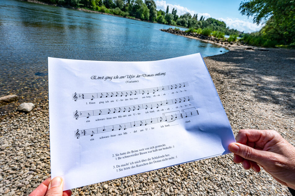 Ein Notenblatt mit dem Liedtext "Einst ging ich am Ufer der Donau entlang" wird am Donauufer gehalten.