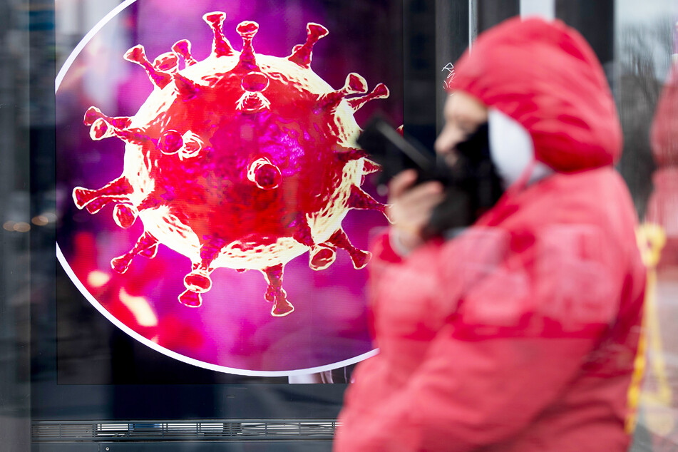 Eine Frau sitzt an einer Bushaltestelle vor einer elektronischen Werbetafel mit einer Illustration des Coronavirus.