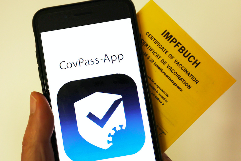Das Logo der CovPass-App ist neben einem Impfbuch auf einem Smartphone zu sehen.