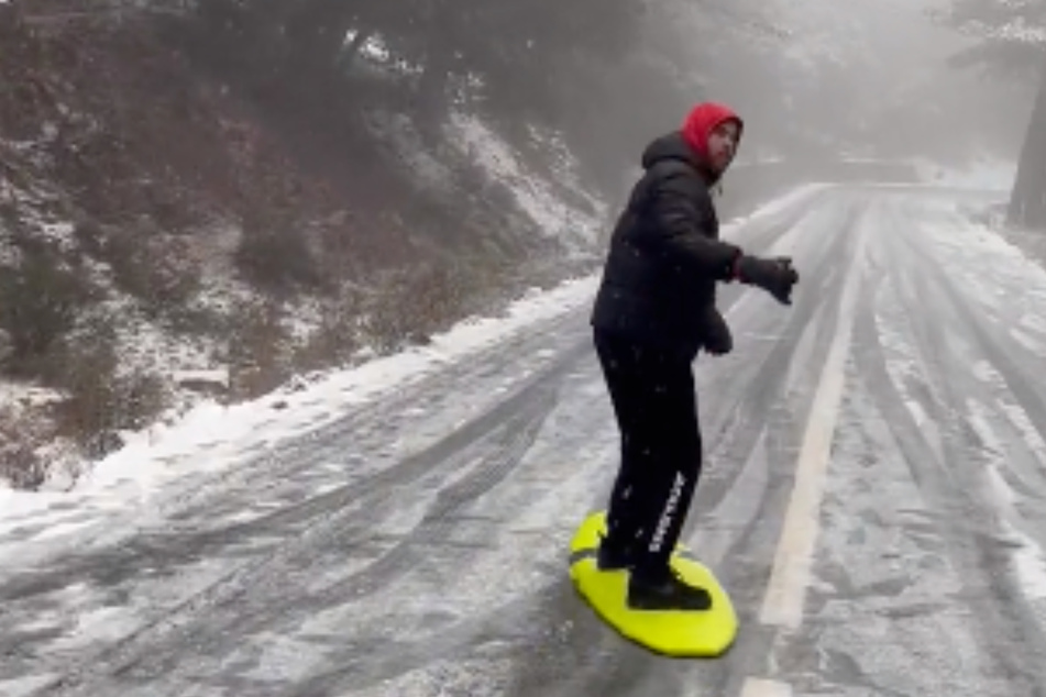 Ein Wagemutiger wandelt sein Surfboard kurzerhand in ein Snowbard um, und fährt damit eine schneebedeckte Straße in den Bergen Mallorcas runter.