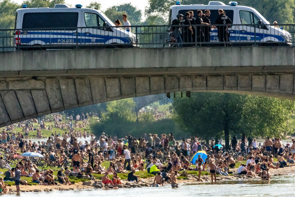 Hunderte Menschen belagern bei sommerlichen Temperaturen die Frühlingsanlagen an der Isar. Auf der Reichenbachbrücke im Vordergrund stehen Einsatzwagen der Polizei.