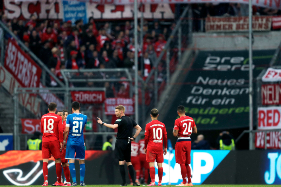 Schiedsrichter Christian Dingert (M) spricht mit Spielern von Hoffenheim und München, während die Münchner Fans auf der Tribüne ein Banner mit der Aufschrift "Hopp bleibt ein Hurensohn" zeigen.