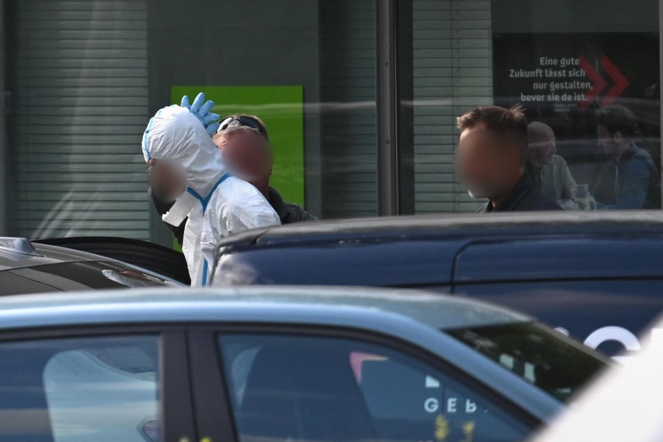 Eine Person in einem speziellen, weißen Anzug wird bei einem Polizeieinsatz an einer Bank weggeführt.