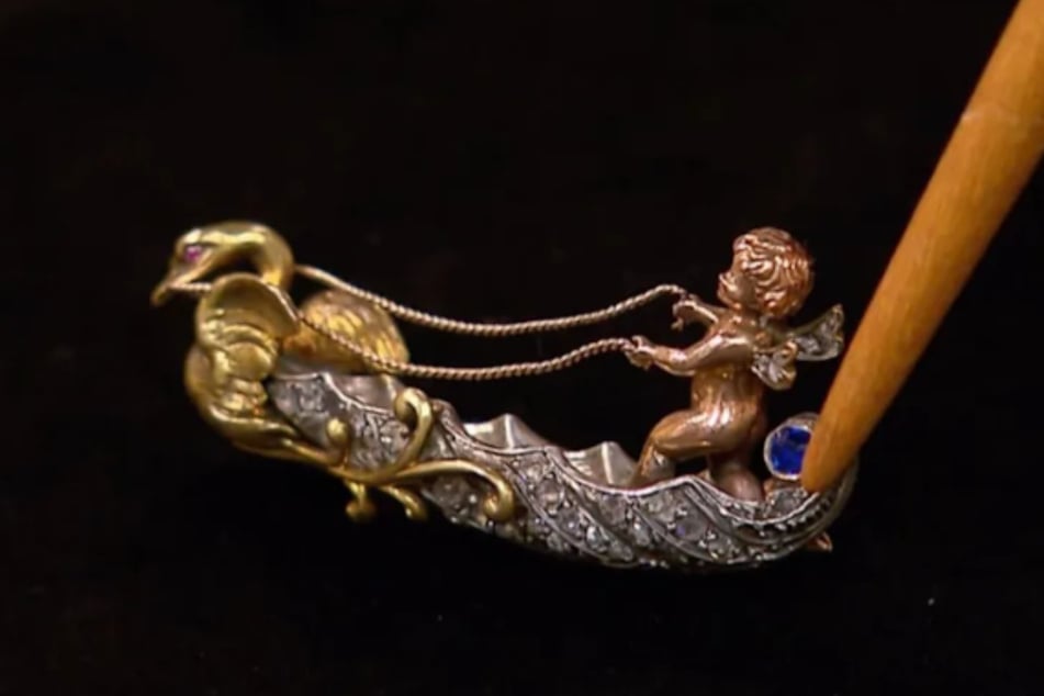 Eine kleine Amorette aus 585er-Rotgold hält die Zügel des Schwans. Das Heck der Barke wird von einem sehr schönen Saphir geziert.
