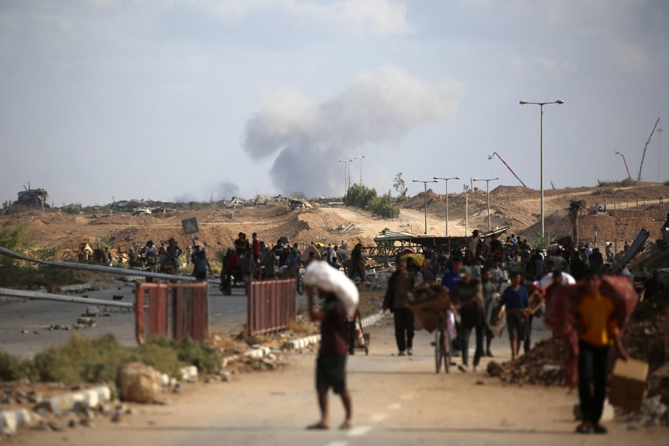 A plume of smoke rises in the background as Palestinians return from a food distribution point run by the US and Israeli-backed Gaza Humanitarian Foundation, near the Netsarim corridor in the central Gaza Strip, on October 5, 2025.