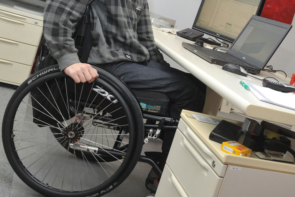 Ein Mann sitzt in einem Rollstuhl an seinem Arbeitsplatz am Schreibtisch.