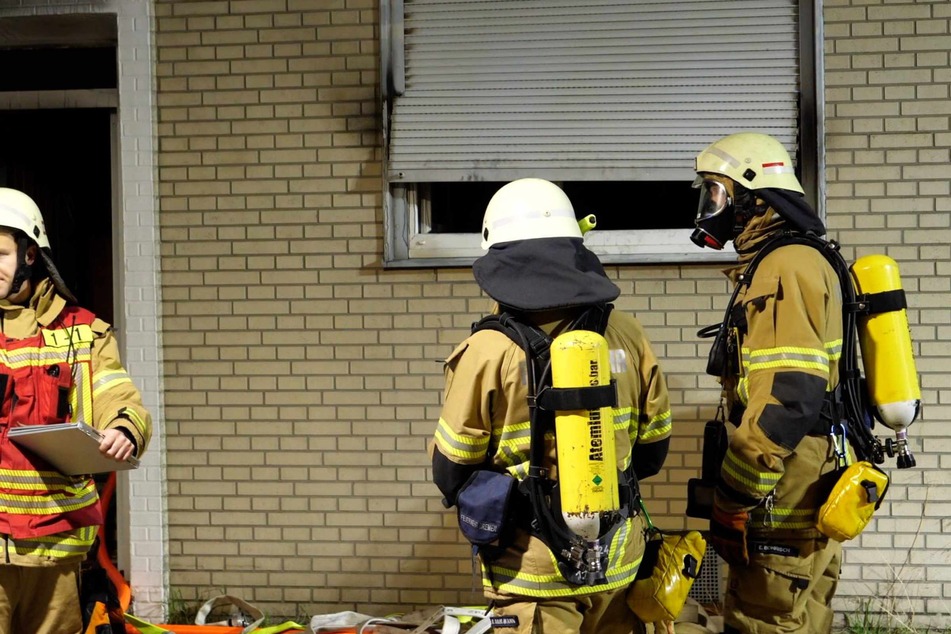 Feuerwehrkräfte stehen vor einem Fenster. Im Hintergrund sind Spuren des Brandes an Fenster und Jalousie zu erkennen.