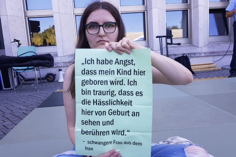 Vor dem Ministerium sitzt eine Protestierende mit einem Schild vor sich.