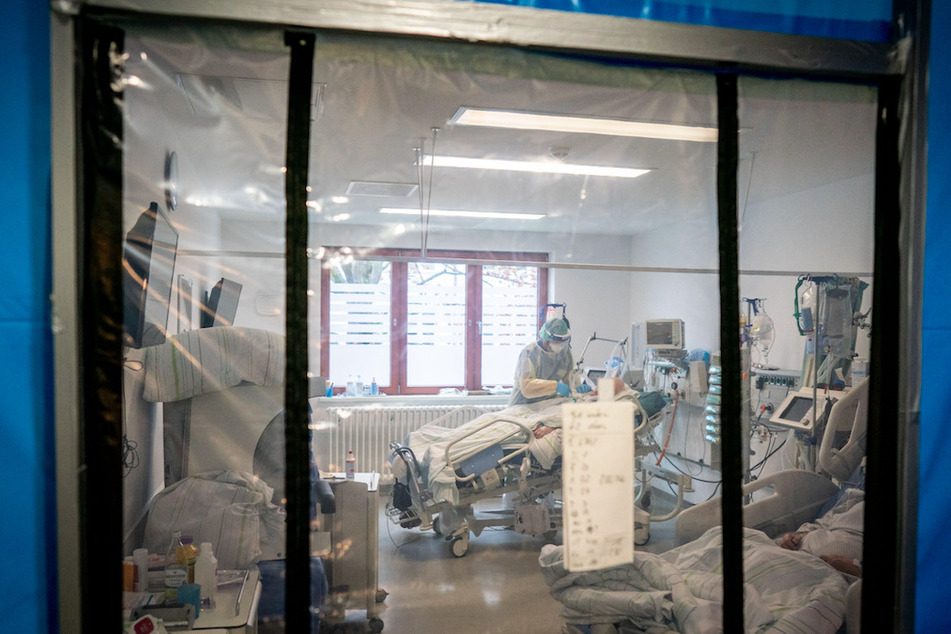 Ein Pfleger ist hinter einer Schleuse bei seiner Arbeit in einer Schutzausrüstung an einem Corona-Patienten zu sehen.