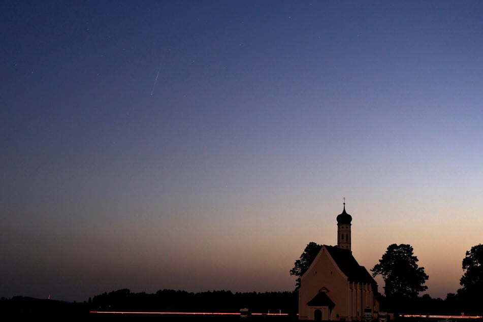 Man muss schon sehr genau hinsehen, um eine kleine Sternschnuppe am Himmel hinter der Wallfahrtskirche Sankt Coloman in Bayern im linken obereren Teil des Fotos zu erblicken.