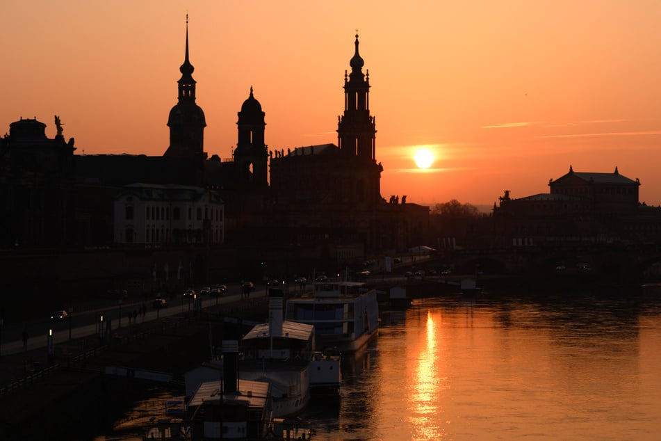 Der Hausmannsturm, das Ständehaus, die Katholische Hofkirche und die Semperoper zeichnen sich vor der untergehenden Sonne am Ufer der Elbe als Silhouetten ab.