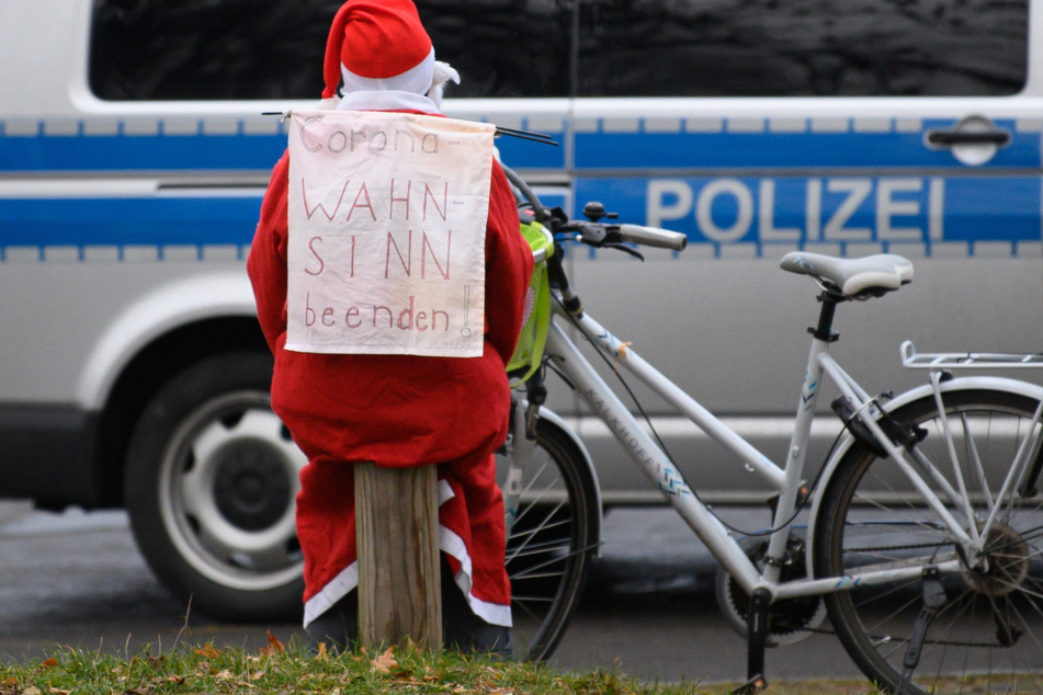 Ein als Weihnachtsmann gekleideter Fahrradfahrer trägt ein Banner "Corona-Wahnsinn beenden!" auf dem Rücken und sitzt an der Cockerwiese auf einem Holzpfahl, während ein Polizeiauto vorbei fährt.