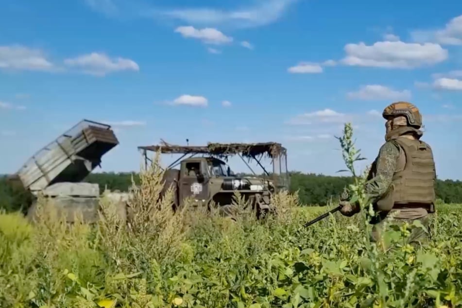Ein russischer Soldat steht auf einem Feld. Derweil macht sich Raketenartillerie bereit, um auf ukrainische Stellungen zu feuern.