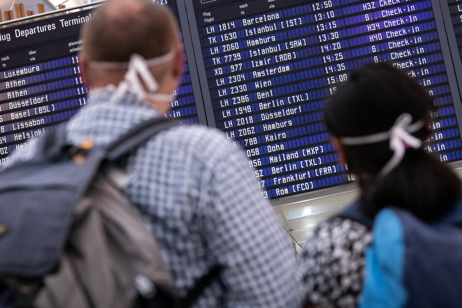 Reisende mit Mundschutz schauen auf eine Anzeigetafel am Münchner Flughafen, auf der zahlreiche internationale Flugziele gelistet sind. (Archivbild)
