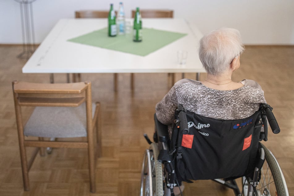 Eine ältere Frau sitzt in einem Pflegeheim in ihrem Rollstuhl vor einem Tisch.