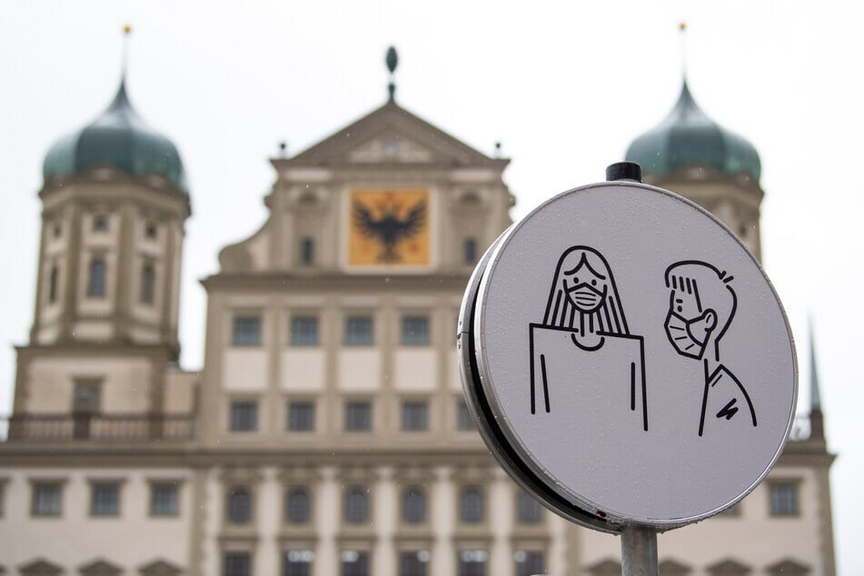 Eine Darstellung der Maskenpflicht ist auf einem Hinweisschild vor dem Rathaus in Augsburg zu sehen.(Archiv)