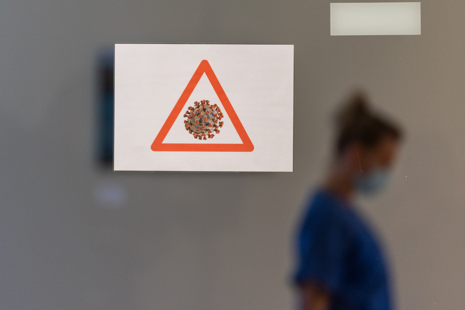 Eine Gesundheits- und Krankenpflegerin läuft auf der Corona-Station im Städtischen Klinikum Dresden in Schutzausrüstung hinter einer Scheibe an der ein Achtungssymbol mit einem stilisierten Cornavirus angebracht ist vorbei.