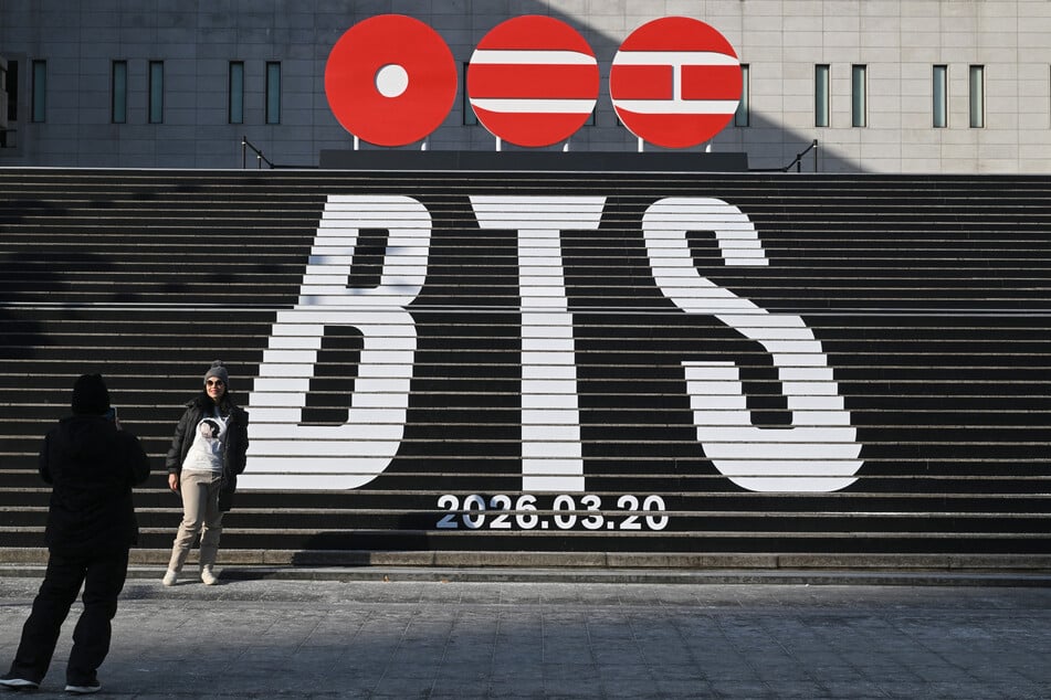 A woman poses for a photo in front of stairs displaying the logo of BTS' 2026 album and release date at Gwanghwamun Square in Seoul on January 14, 2026.