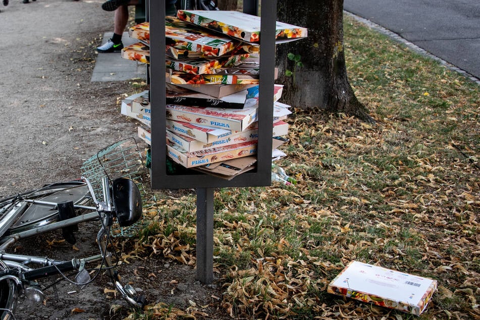 Pizzakartons sind in einem Stahlgestänge für einen Mülleimer in einer Parkanlage gestapelt und liegen teilweise auf dem Rasen.