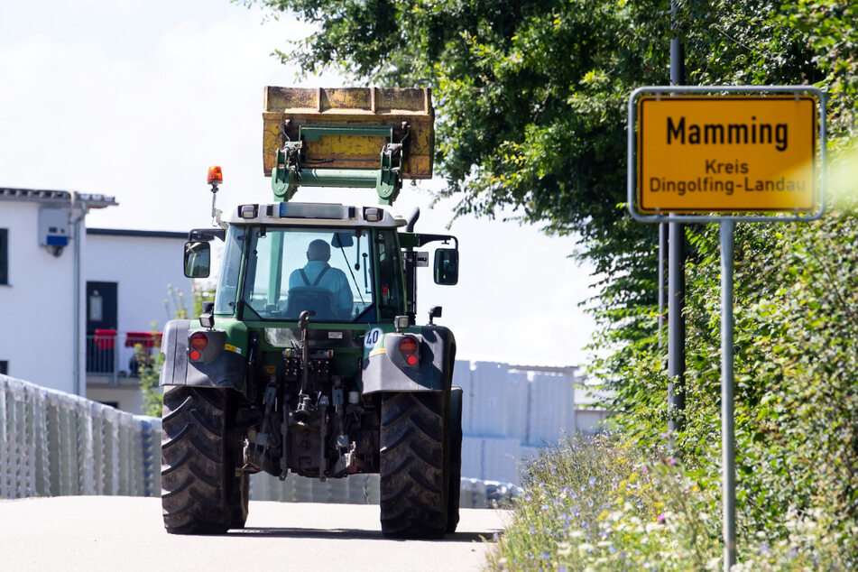 Ein Landwirt fährt mit seinem Traktor am Ortsschild von Mamming und einer Konservenfabrik entlang.