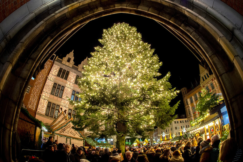 Besucher gehen an einem mit Lichtern geschmückten Tannenbaum auf dem Weihnachtsmarkt vorbei.