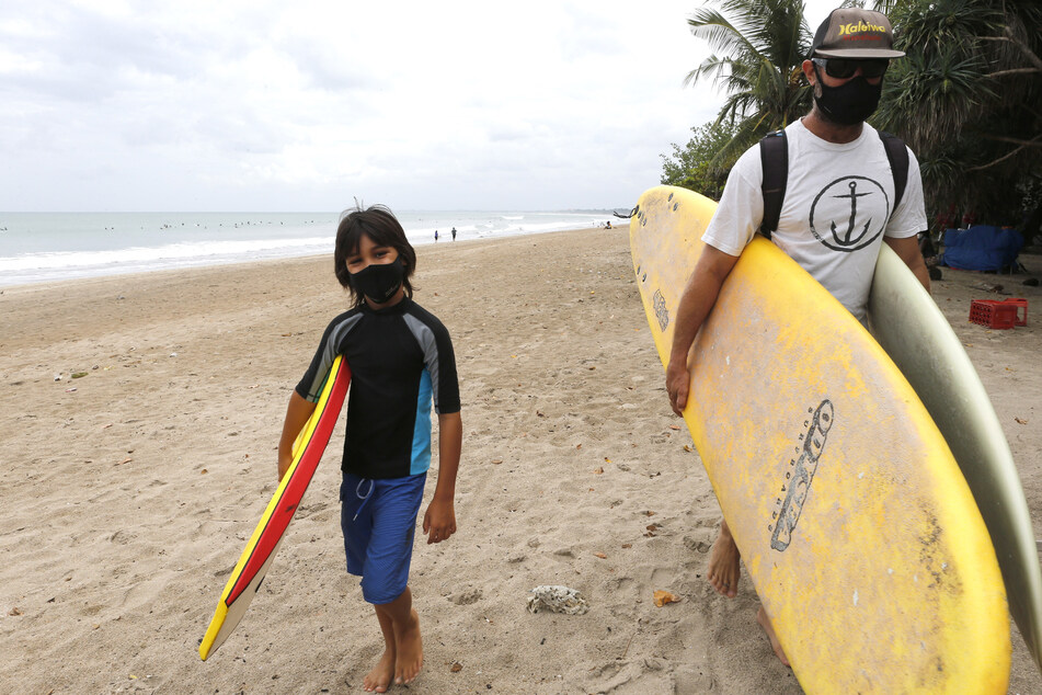 Touristen mit Mundschutz tragen ihre Surfbretter am Strand von Kuta.