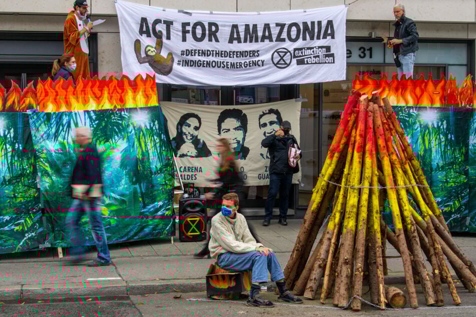 Ein Demonstrant ist auf einem mit symbolischen Flammen verzierten Holzklotz an einem großen symbolischen Feuer festgekettet.