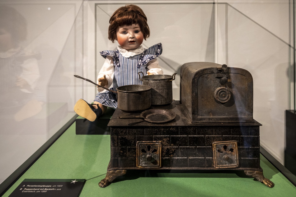 Eine Porzellankopfpuppe (um 1920) steht hinter einem Puppenherd mit Backofen (um 1890).