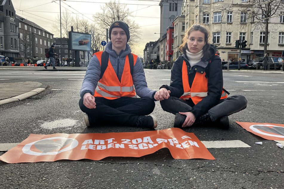 Zwei Aktivisten der "Letzten Generation" sitzen Hand in Hand für mehr Klimaschutz auf der Straße.