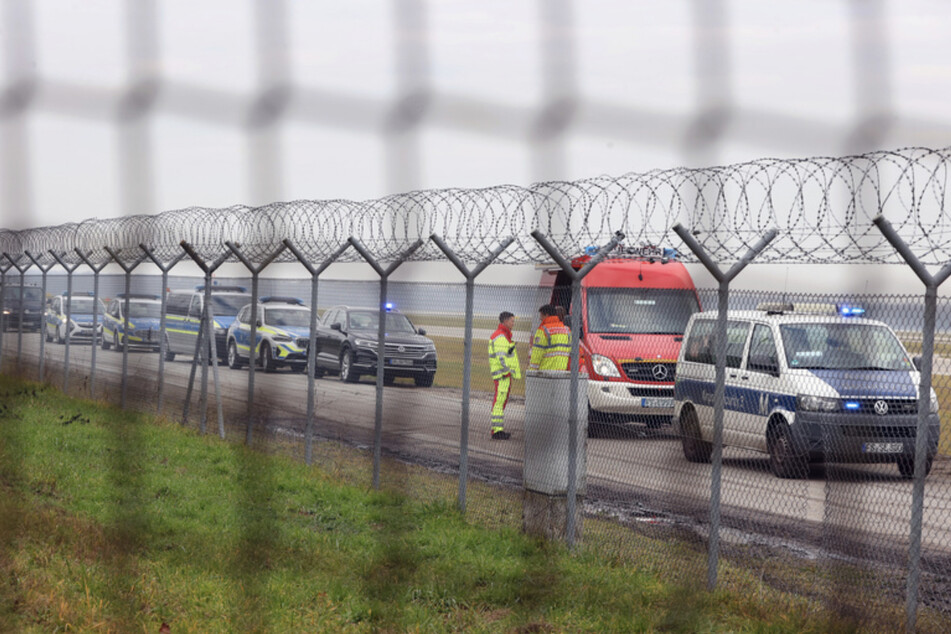 Einsatzkräfte von Polizei und Feuerwehr stehen neben dem Zubringer einer Start- und Landebahn am Airport Franz-Josef-Strauß, auf dem sich vier Klimaaktivisten angeklebt haben.