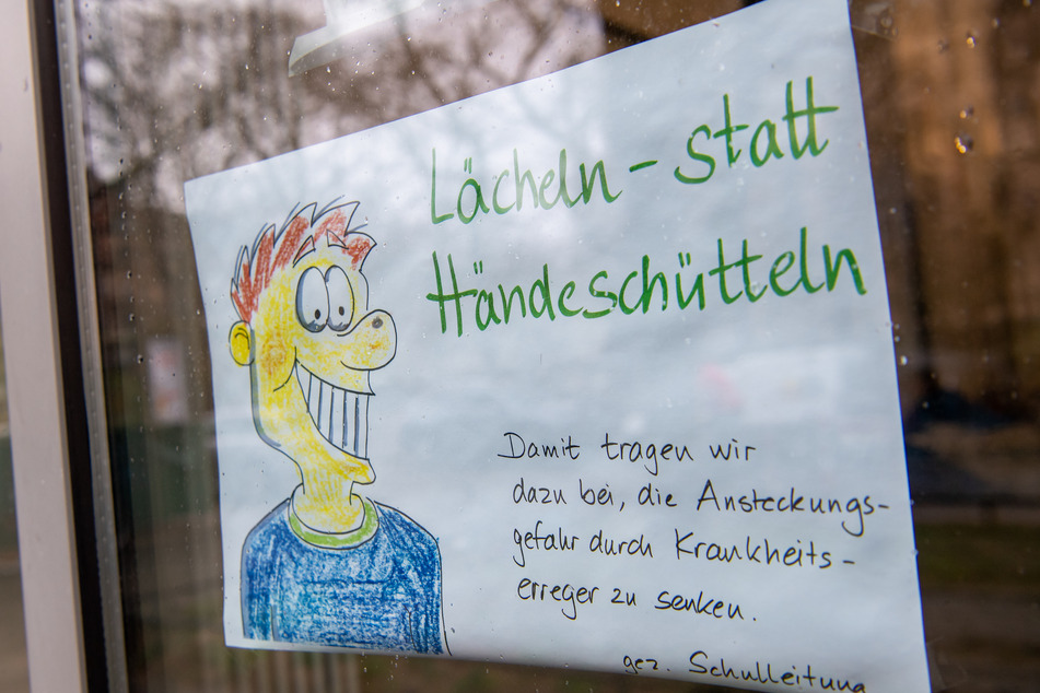 Ein gemaltes Schild mit der Aufschrift "Lächeln statt Händeschütteln- Damit tragen wir dazu bei, die Ansteckungsgefahrt durch Krankheitserreger zu senken - gez. Schulleitung" hängt an der Tür eines Grundschule.