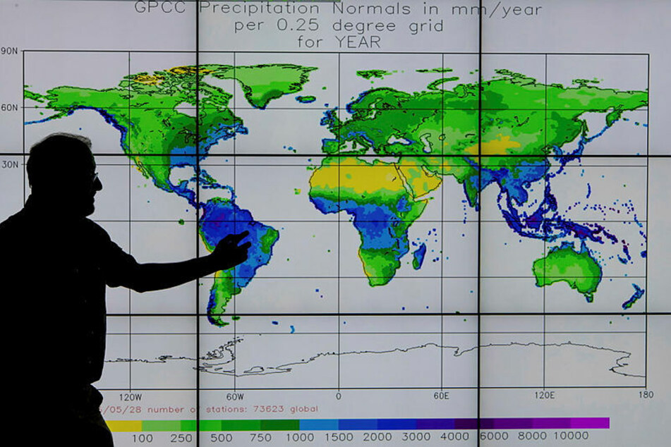 Nur als Schattenriss ist dieser Meteorologe zu erkennen, der vor einer Bildschirmwand steht, auf der die weltweiten Niederschlagsmengen farbig dargestellt werden.
