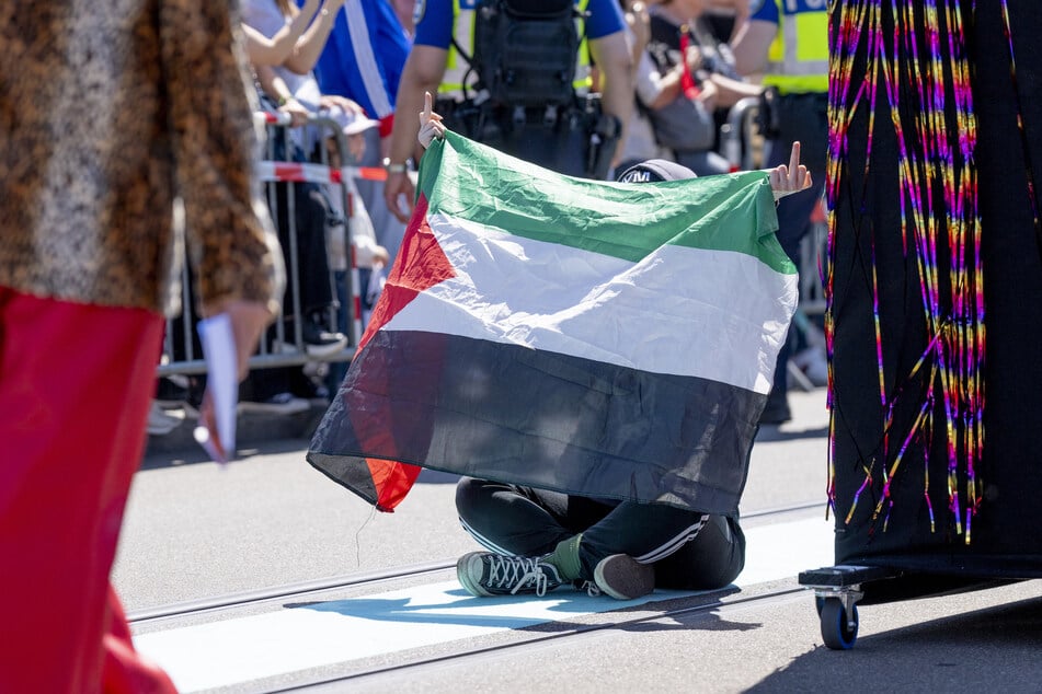 Ein Demonstrant sitzt bei einer ESC-Veranstaltung in Basel mit Palästina-Flagge auf dem Bode, zeigt seine Mittelfinger.